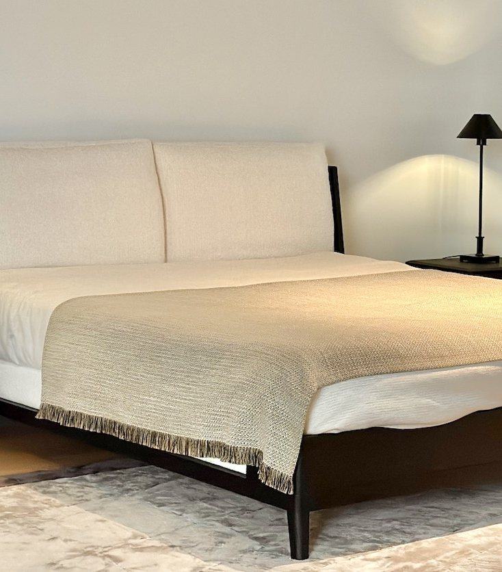 A dark timber bed with a soft ivory headboard cushion and a woven throw